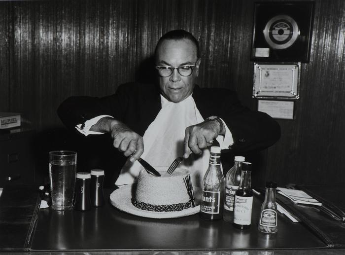 A man cutting a cake shaped like a hat.