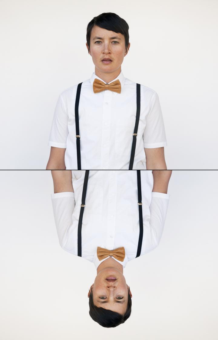 An individual's profile staring at the camera and their mirror image reversed, wearing a brown bowtie and suspenders. 